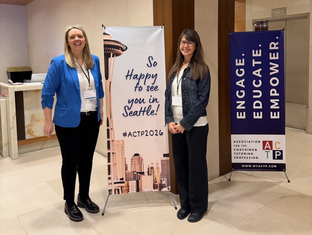 Two women smiling beside banners. Left banner says "So Happy to see you in Seattle! #ACTP2026" with the Space Needle. Right banner reads "Engage. Educate. Empower."