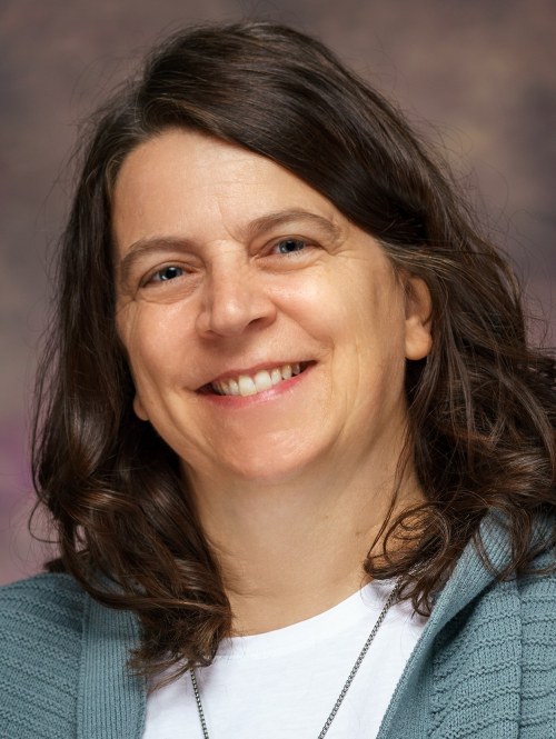 Close-up portrait of a smiling woman with shoulder-length brown hair, wearing a blue cardigan over a white shirt and a necklace.