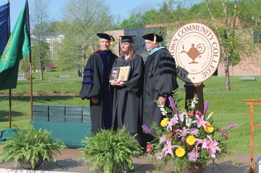 Three people in caps and gowns, one holding a plaque, stand on an outdoor stage during a commencement ceremony.