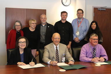 Three people sit at a table with papers in front of them that they are signing while six more people stand behind them, smiling.