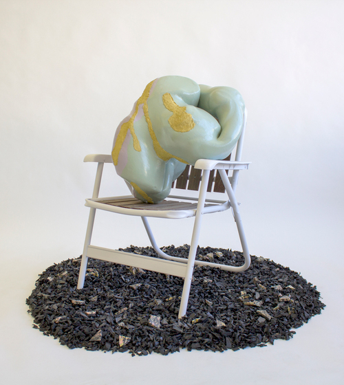 A milky green sculpture with gold accents placed on a vintage lawn chair rests in material reminiscent of dark mulch.