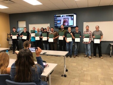 A group of program graduates stand holding their certificates while onlookers, seated at tables, take pictures with their phones.