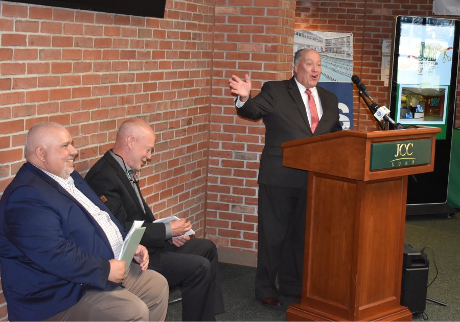 A man in a suit gestures while speaking from behind a podium with "SUNY JCC" signage, as two other men sit attentively against a brick wall.