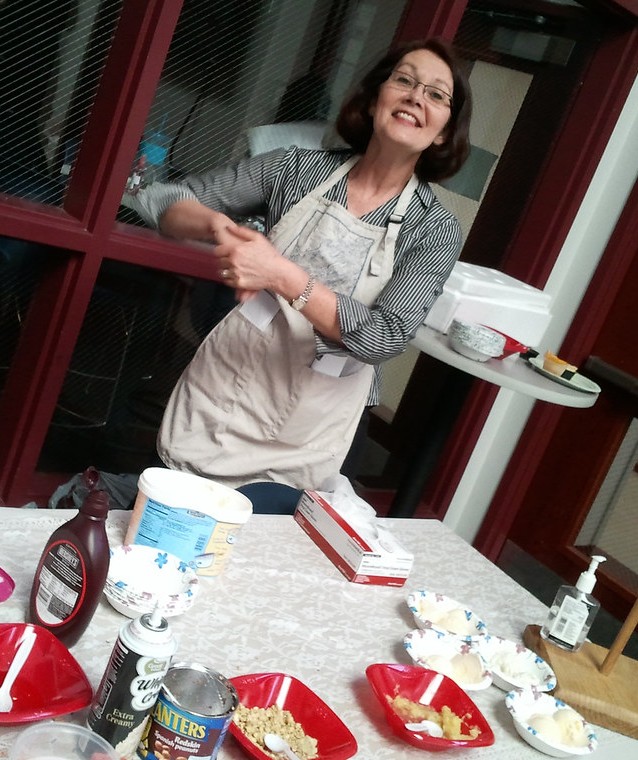 Smiling woman in glasses and apron prepares food in a kitchen, surrounded by baking supplies, dishes, condiments, and a serving tray.