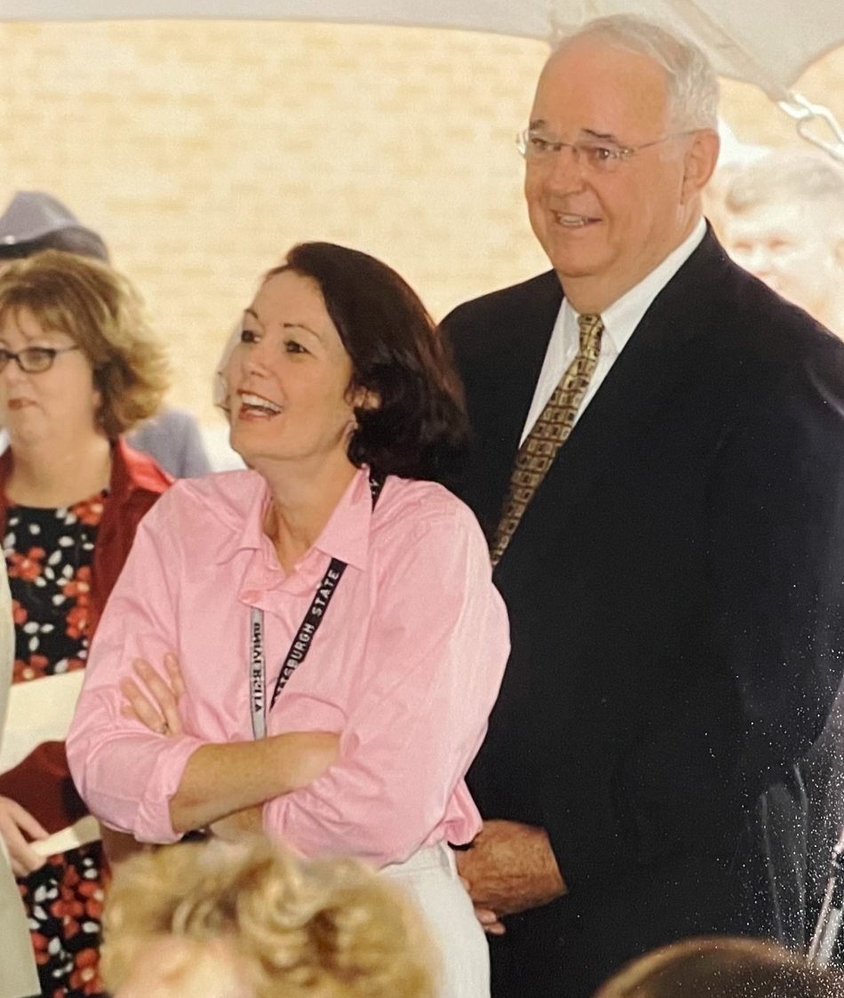 Adults, including a smiling woman in pink and an older man in a suit, gather under a tent at a cheerful outdoor event.