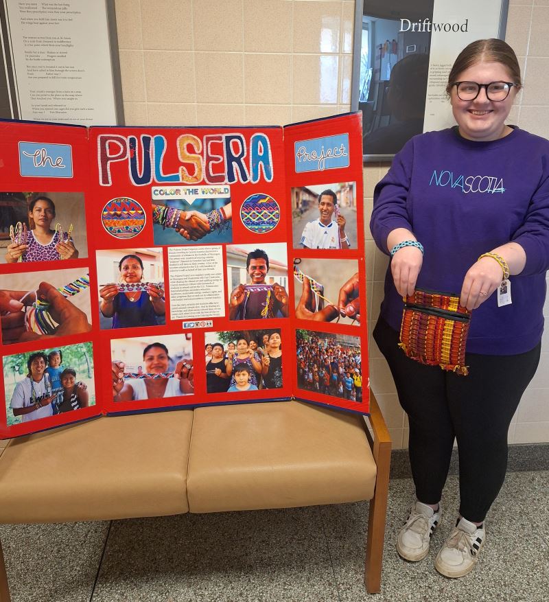 A person wearing handmade bracelets and holding a handemade purse poses near a presentation board that shows information and photos from the Pulsera Project.