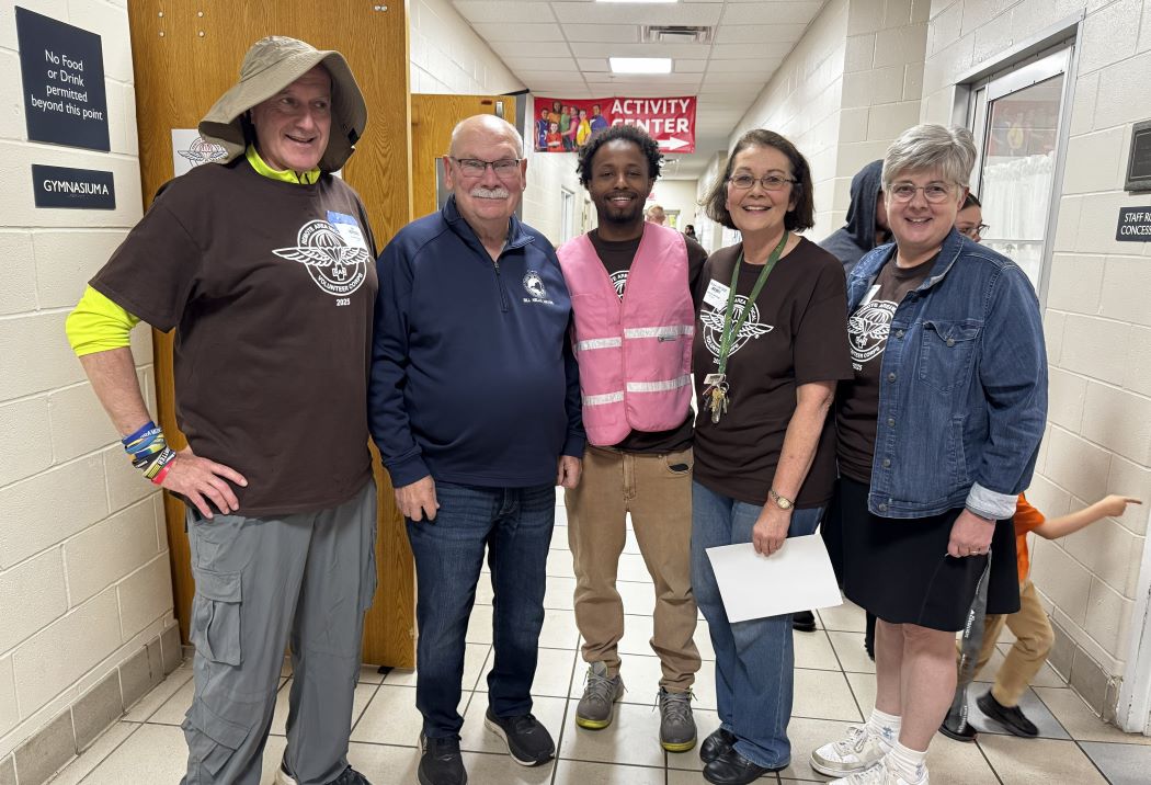 Five people stand in a hallway smiling. Three wear brown shirts, one a pink vest, and another a denim jacket.