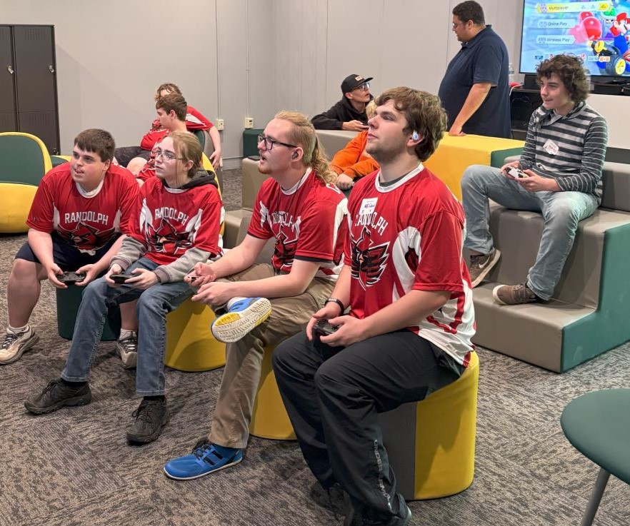 Students in red Randolph uniforms hold gaming controllers while playing a video game.