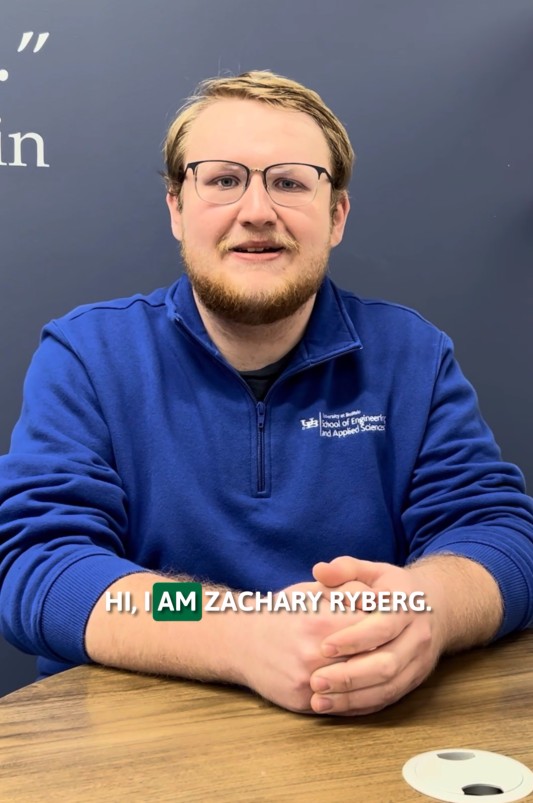 Man with glasses and beard in blue School of Engineering sweater, seated at a table against a navy blue wall, smiling, with text overlay that reads Hi, I am Zachary Ryberg.