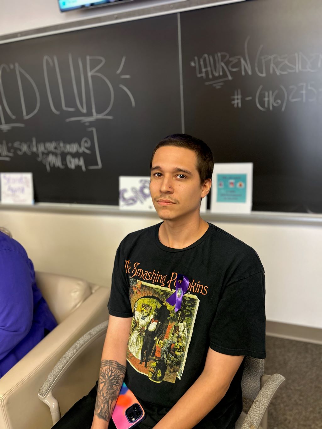 A person in a college classroom sitting at the front of the room with a chalkboard in the background.