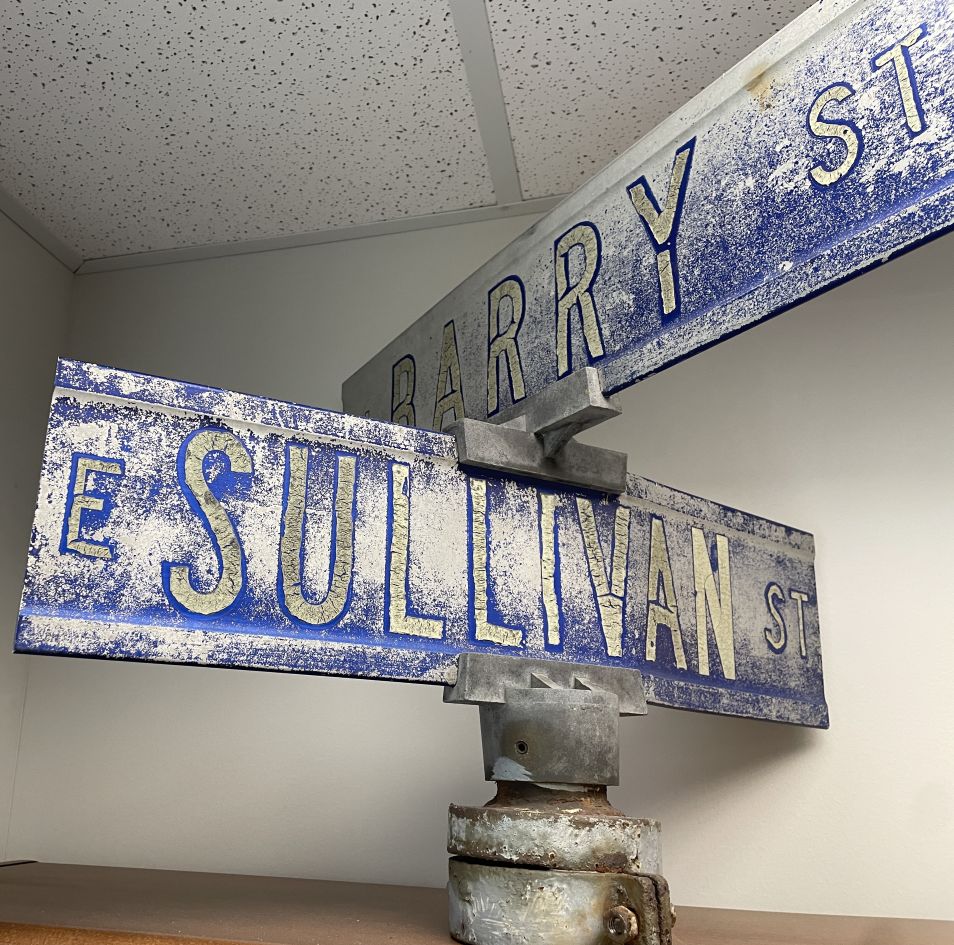 Weathered blue-and-white metal signpost reading “SULLIVAN ST” and “BARRY ST,” mounted indoors with rusty hardware and an industrial feel.