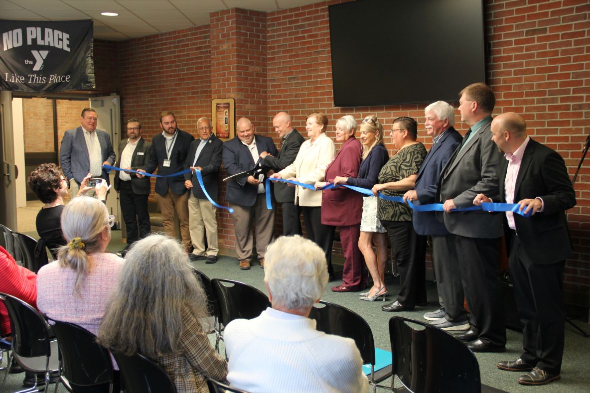 A group of people stand in front of a brick wall, holding a blue ribbon stretched across the image. Several individuals are in suits, and some are holding scissors, preparing to cut the ribbon. A banner with the YMCA logo and the words "NO PLACE Like This Place" is visible on the left side of the image. An audience sits in chairs facing the ribbon-cutting ceremony.