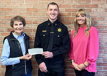 A woman stands to the left of a man in a police officer's uniform. They are holding a check and there is a woman standing with them to the right smiling with her hands folded.