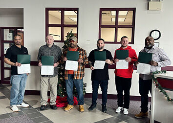 Six people stand holding their program graduation certificates in a room with two windows behind them.