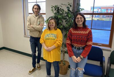 Three students pose together with a fake plant and windows in the background.