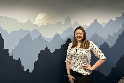 An artist stands in front of a painted mural of mountains, a moon and sky on an interior wall.