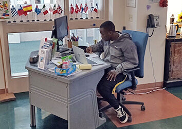 A student worker sits at a desk in front of windows with small international flags lined up on the ledge behind him.