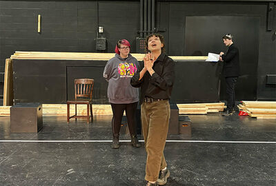 Actors practicing on a stage with a wooden chair in the background and stacks of unfinished boards behind the chair.