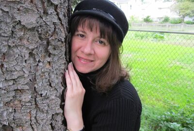 A person wearing a hat leans with their hands against a tree with a chain-link fence and green grass in the background.