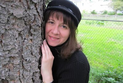 A person wearing a hat leans with their hands against a tree with a chain-link fence and green grass in the background.