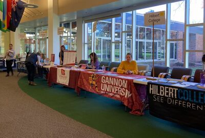 Representatives from college sit at tables in a student union with table cloths showing their college logos.