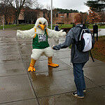A hawk mascot in a JCC jersey with arms outstretched offering a hug to a person wearing a jacket, knit cap and backpack.