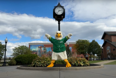 The JCC Jayhawk mascot leaps into the air on campus with a clock tower and building in the background.