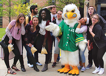 A bird mascot in a button-down shirt stands amongst a group of students posing in various ways.