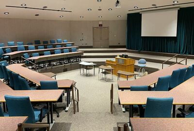 A look inside an academic theater with rows of seating and chairs, a podium, and large projector screen.