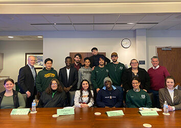 A row of people sitting at a table in a board room with name cards that say "Evelyn Burdick, Julianna Main, Ayomideji Israel-Akinbo, and Bethan Wallace" with a row of people standing behind them. 