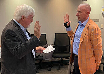 Two men mirroring each other with one hand raised while one reads an oath from a book during a swearing in ceremony.