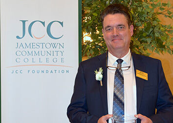 Scott Kindberg wearing a suit and tie standing in front of a tall banner that says "JCC Jamestown Community College SUNY JCC Foundation."