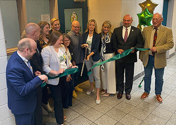 A group of people stand together while the two at the outer edges hold a ribbon and a woman in the middle cuts it with a giant pair of scissors.