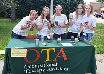 Five students are holding jars filled with glittery liquid and standing behind a table on a sidwalk with a tablecloth that says OTA Occupational Therapy Assistant.