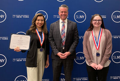 Three people stand in front of a State University of New York backdrop. The first on the left is holding a certificate and wearing a medal, and the third from left is also wearing a medal.