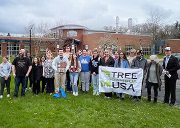 A group of people stand together in front of a brick building and some are holding a banner that says "Tree Campus USA."
