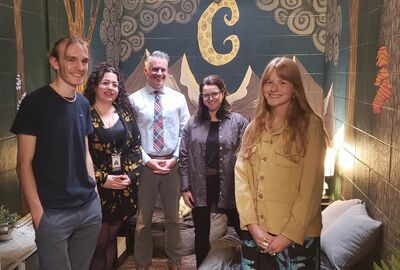 A group of five smiling people pose together in a college campus meditation room featuring a rainforest-themed mural on the walls.