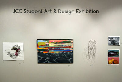 Student paintings and drawings are displayed on an art gallery wall that also includes the words JCC Student Art and Design Exhibition.