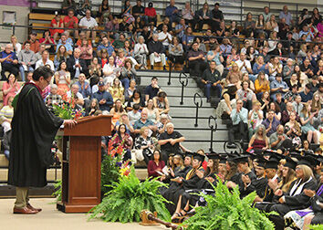 A man stands at a podium on stage in a graduation gown while speaking to a gymnasium full of people in the bleachers and in chairs on the floor in front of him.