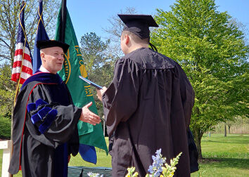 People in ceremonial graduation robes stand outside while exchanging a degree during an outdoor commencement ceremony.