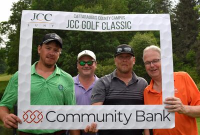Four golfers pose for a photo while holding up and standing behind a white golf ball textured frame that reads Cattaraugus County JCC Golf Classic and Community Bank.