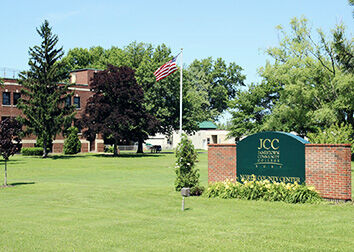 The front lawn of SUNY JCC North County Center with sign in a brick frame, American Flag, and trees.