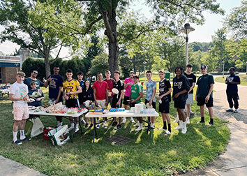 People standing around a table full of food outside on a grassy lawn framed by sidewalks. 