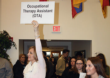 A woman holds a sign that says "Occupational Therapy Assistant (OTA)" in the entrance of a room with people behind and beside her.