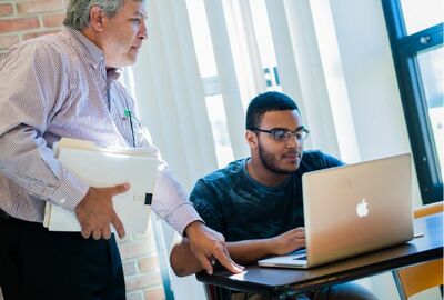 A student looks at laptop computer while a college professor stands nearby looking over his shoulder.