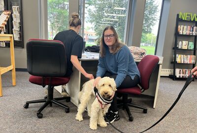 Two students pet a therapy dog while sitting at a study table in a college library.