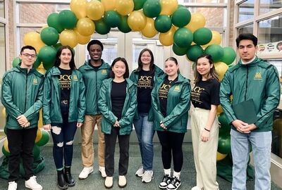 Students dressed in green JCC logo jackets stand in front of a green and gold balloon archway.