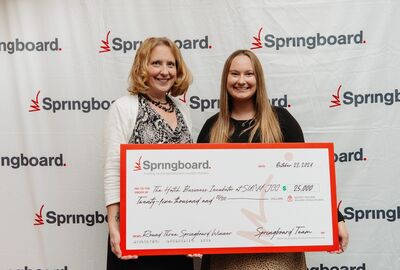 Two people smile while holding a large check worth $25,000 in front of white background with many Springboard logos on it.