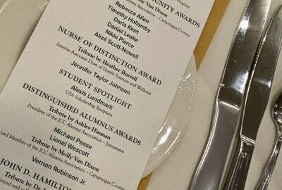 A dinner awards gala agenda laying on a dinner plate with silverware nearby on a tablecloth.