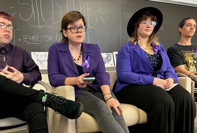 Four students seated in chairs in the front of a classroom with a black chalkboard in the background.